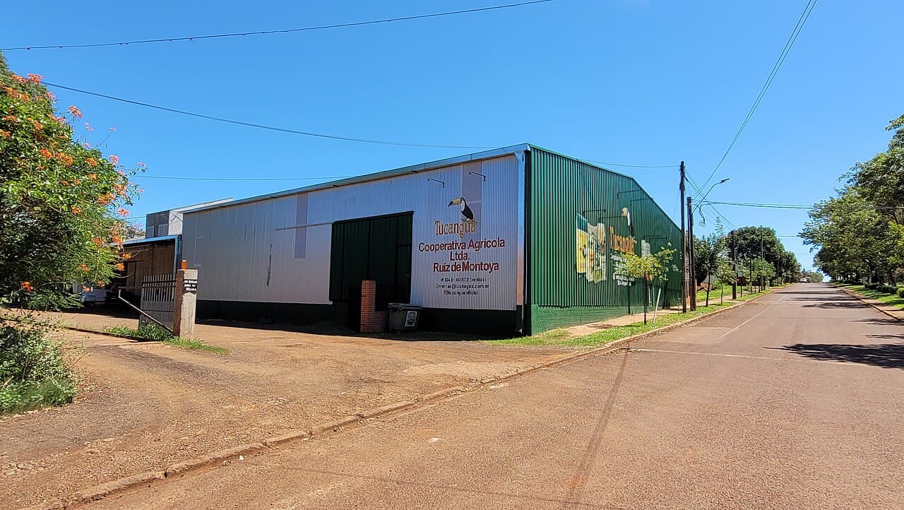 The green-and-white corrugated-steel premises of the Cooperativa Agrícola Ltda. Ruiz de Montoya on Avenida de los Inmigrantes in Misiones, Argentina, the agricultural cooperative's name and its 'Tucanguá' toucan brand painted across the warehouse wall beside a quiet red-earth street