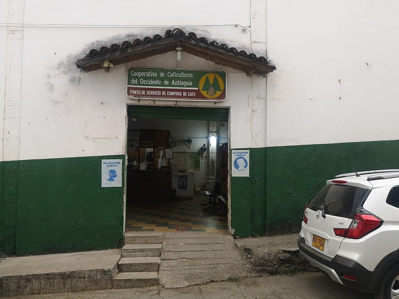 A coffee-buying point of the Cooperativa de Caficultores del Occidente de Antioquia at Sopetrán, Colombia — a whitewashed building with a green dado, its tiled porch carrying the coffee cooperative's sign reading 'Punto de Servicio de Compras de Café' above the open doorway