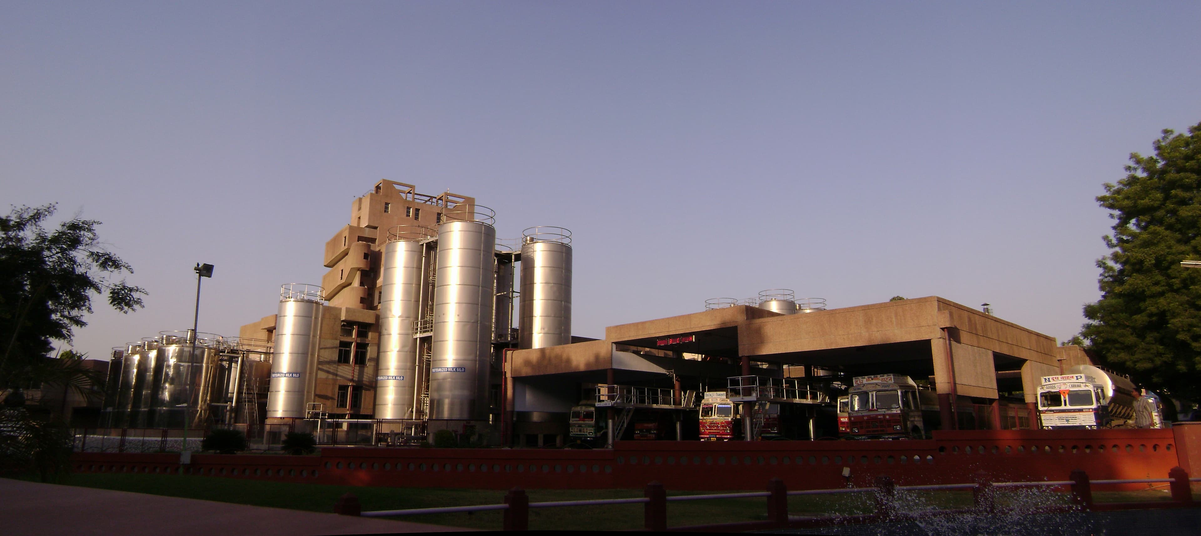 The Amul dairy cooperative processing plant at Anand, Gujarat, at dusk — tall stainless-steel pasteurised-milk silos beside the concrete processing building, with milk tanker trucks lined up at the loading bay