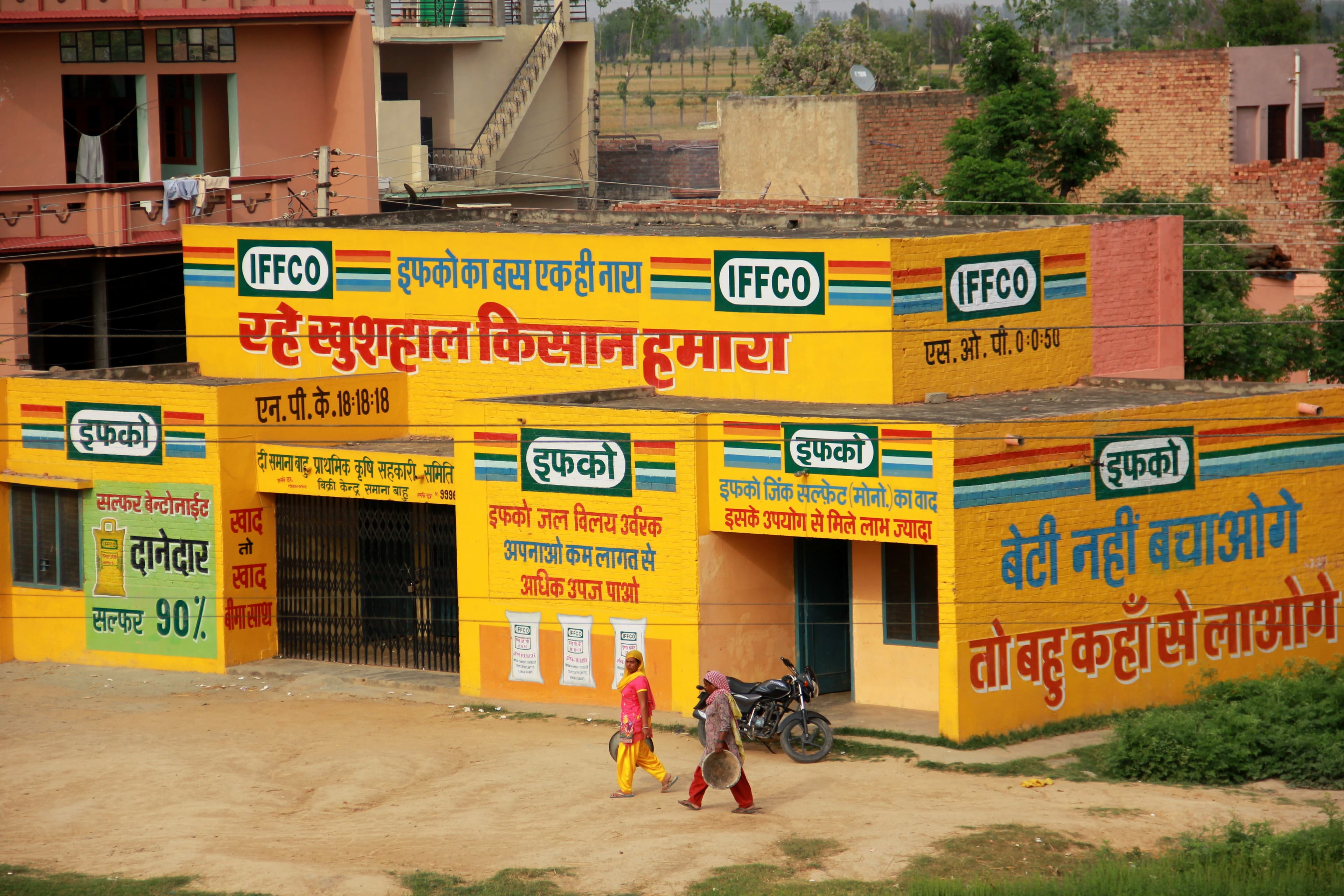 A village IFFCO fertiliser-cooperative sales point in rural India — a bright-yellow primary agricultural cooperative society building painted with IFFCO branding and crop-input slogans in Hindi, two women in saris walking past on the dirt forecourt