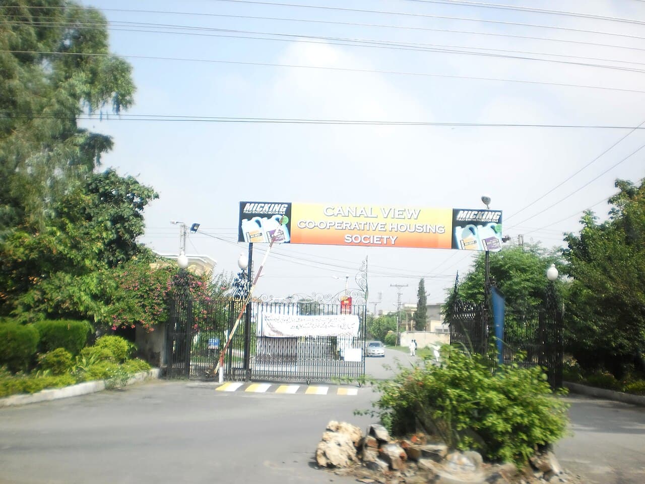 The gated entrance of the Canal View Co-operative Housing Society in Lahore, Pakistan — a wide overhead signboard reading 'Canal View Co-operative Housing Society' spanning the road above ornamental black railings and a landscaped median
