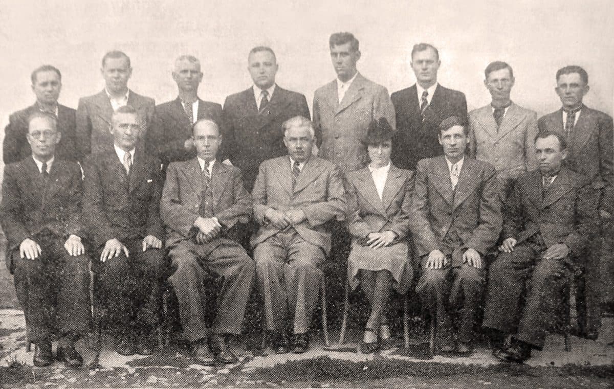 The directors and staff of the Buchach District Union of Cooperatives in western Ukraine, photographed in 1934 — a formal black-and-white group portrait of fifteen cooperators in suits, fourteen of them named in the Commons record and one unidentified, seven seated in front including one woman, eight standing behind, against a pale wall